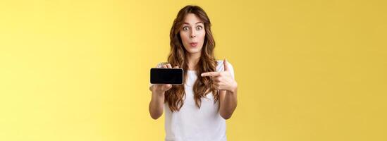 Excited surprised pleased girl curly long hairstyle folding lips whistling amused stare camera impressed showing smartphone pointing index finger mobile cellphone screen stand yellow background photo