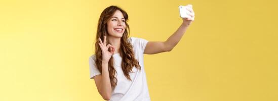 Stylish carefree urban confident curly-haired girl recording video blog followers extend arm hold smartphone taking selfie front camera show okay ok approval gesture yellow background photo