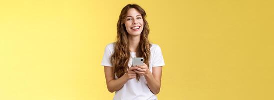 Lively enthusiastic friendly smiling happy woman using smartphone texting messaging friend checking social media feed browsing internet hold mobile phone laughing happily yellow background photo