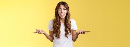 Pissed frustrated bothered curly-haired caucasian girl stare camera puzzled complain hold smartphone shrugging spread hands sideways dismay arguing after intense serious phonecall yellow background photo