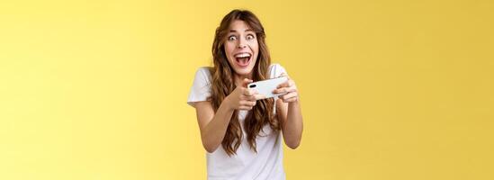 Extremely happy thrilled playful girl gamer playing awesome great new smartphone game hold mobile phone horizontal cheering look camera astonished impressed beating record yellow background photo