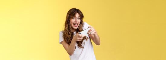 Excited playful enthusiastic girl tilting sideways playing awesome interesting smartphone game car racing smiling determined focuse gaming hold mobile phone horizontal tap display yellow background photo