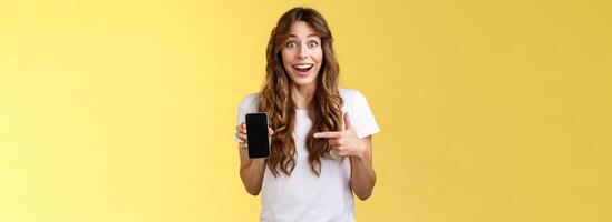 Surprised happy lucky girl winning online internet lottery smiling broadly hold smartphone pointing blank mobile phone screen showing display grinning excited enthusiastic stand yellow background photo