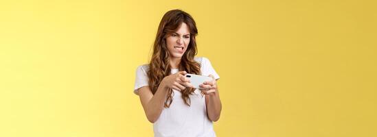 Excited focused playful enthusiastic geeky attractive girl trying beat score hold smartphone horizontal grimacing intense look screen playing awesome arcade game yellow background photo