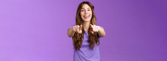 Its you. Friendly glad joyful happy curly-haired girl pointing fingers camera smiling broadly laughing congratulate friend singing song indicating boyfriend make choice stand purple background photo