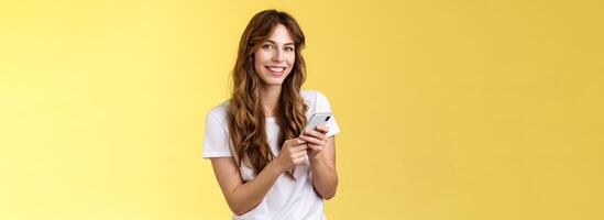 Enthusiastic good-looking urban girl wear white t-shirt standing casual smiling delighted camera texting hold smartphone scroll social media feed stand yellow background browsing network photo