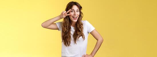 Playful surprised charismatic young caucasian girlfriend having fun look amused enthusiastic show peace victory sign laughing smiling joyfully enjoy summer holidays touch hip stand yellow background photo