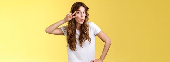 Cute glamour young woman curly haircut tilt head silly show peace victory sign fold lips lovely muah kiss gesture send flirty glances camera hold hip mimicking sensually gazing you yellow background photo