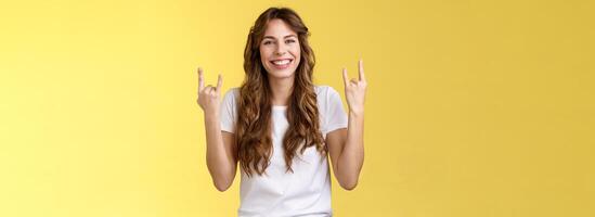 Happy cheerful feminine caucasian girl curly hairstyle show rock-n-roll heavy metal gesture smiling broadly enjoy awesome concert atmosphere stand yellow background joyfully cheering satisfied photo