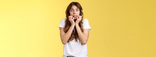 Scared timid insecure unconfident european girl curly haircut hold hands face biting fingers look frightened shocked standing stupor speechless shaking fear terrified yellow background photo