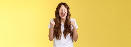 Excited happy triumphing joyful caucasian girl close eyes fist pump celebratory happiness gesture yelling yeah success reach goal achievement dancing victory winning feel relieved yellow background photo