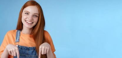 Friendly happy redhead girl smiling lively pretty grin pointing down index fingers offering good offer recommend use service standing blue background discuss interesting product, blue background photo