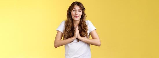 Cute hopeful young caucasian girl stooping hold hands pray smiling hope for pity begging favour plead asking advice say sorry grinning lovely making faithful glances you lend hand yellow background photo