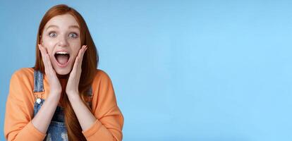 Excited thrilled young emotional enthusiasitc ginger girl teenage college student yelling amused smiling broadly receive positive good news look surprised camera touch face astonished blue background photo