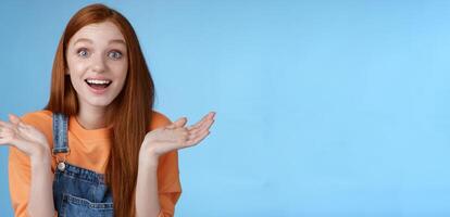 Amused surprised glad young redhead girl speechless happy see friend came back hometown wide eyes impressed grinning raise hands sideways full disbelief standing blue background joyful photo