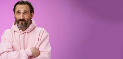 Unsure troubled caucasian bearded adult guy with earring pink hoodie press lips frowning counting mind standing hesitant doubtful thinking look up uncertain taking important decision photo