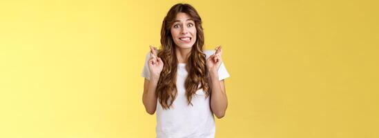 Excited worried cute silly girlfriend anticipating important results nervously biting lower lip unconfidently cross fingers good luck smiling hope dream come true making wish yellow background photo