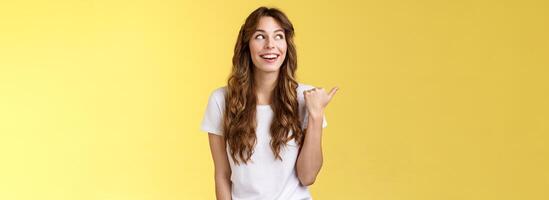 Something interesting right there. Curious lively enthusiastic charming woman glancing pointing thumb left smiling broadly discuss cool event laughing carefree stand yellow background photo