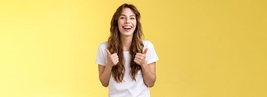 Pleased outgoing cheerful good-looking caucasian girl curly long haircut white t-shirt show thumbs up smiling lively pleased like awesome performance approve good choice yellow background photo