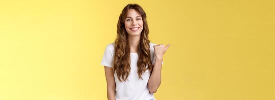 Visit see yourself. Cheerful charismatic good-looking outgoing girl long curly haircut showing place do good hairstyle smiling happily delighted pointing thumb left introduce promo yellow background photo