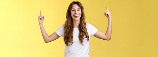 Cheerful amused happy lively young girl long curly haircut raise hands pointing up smiling toothy happily camera introduce excellent variant suggest you click site link advertising yellow background photo