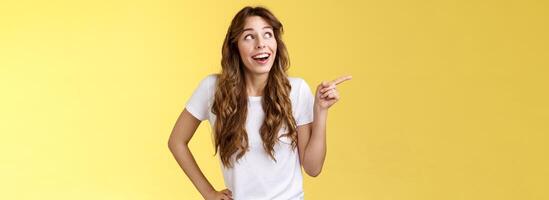 Wondered impressed charismatic fascinated smiling happy girl pointing look upper left corner speechless surprised grinning toothy happiness joy expression contemplate great view yellow background photo