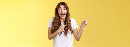 Happy sincere cheerful young surprised woman see celebrity lose speech stare excited unbelievable awesome luck fascinated pointing index fingers left open mouth stunned thrilled yellow background photo