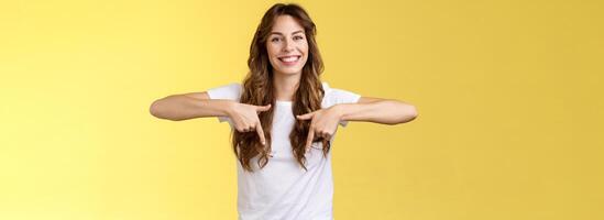 Lively helpful good-looking caucasian girl curly long hair showing you excellent place find hairdresser pointing down index fingers smiling toothy happy delighted grin stand yellow background photo