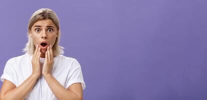 Waist-up shot of worried and shocked woman expressing empathy frowning holding hands near opened mouth feeling sorry and nervous hearing terrible disaster happened standing over purple wall photo