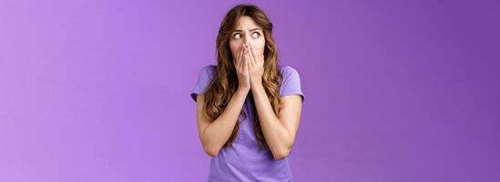 Shocked timid insecure concerned curly-haired woman look sideways stunned scared gasping cover mouth hold palms pressed face stare left frightened troubled panic purple background photo