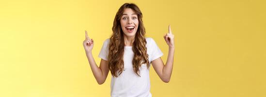 Surprised cheerful celebrating girl lively discuss near awesome event pointing up index fingers smiling broadly fascinated look temtation admire great concert stand yellow background impressed photo