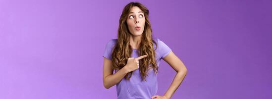 Intrigued lively attractive feminine curly-haired female pointing looking left impressed folding lips say wow astonished express admiration enthusiastic check out awesome promo purple background photo