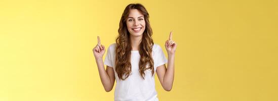 Self-assured confident lively charming european tall girl showing advicing cool promo pointing up index fingers smiling toothy delighted introuce excellent choice advertisement yellow background photo