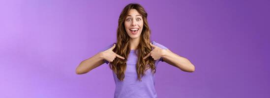 Cheerful lively upbeat entertained happy girl curly hairstyle open mouth amused pointing center herself happily tell own achievement smiling broadly telling friends promotion purple background photo