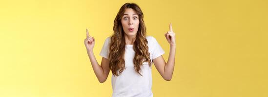 Impressed enthusiastic surprised ambushed cute female model fold lips wow admiration joy stare camera fascinated pointing up index fingers top copy space show you stunning offer yellow background photo