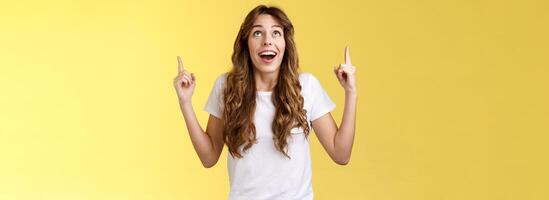 Happy impressed attractive girl long curly haircut drop jaw amused smiling surprised delighted look pointing up index fingers stare admiration fascinated awesome promo offer yellow background photo