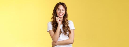 Intrigued attractive european brunette curly long hair look interested listen curious suggestion touch cheek interest temptation smiling toothy pleased good conversation stand yellow background photo