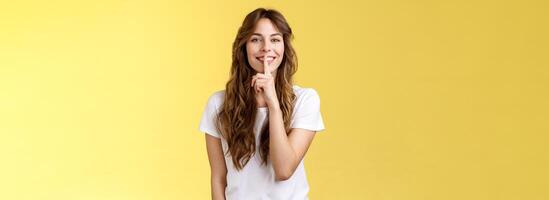 Cute cunning lovely european girl curly hairstyle hiding beauty secret smiling sensually show hush shush gesture index finger pressed lips grinning joyfully stand yellow background photo