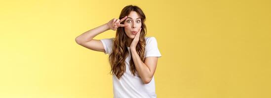 Silly coquesttish good-looking caucasian girl curly hairstyle folding lips amused interested touch cheek feminine sensual pose show victory peace sign upbeat stand intrigued yellow background photo