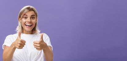 Waist-up shot of optimistic and supportive good-looking caucasian female in white t-shirt cheering and smiling broadly hearing awesome idea approving it with thumbs up gesture over purple wall photo