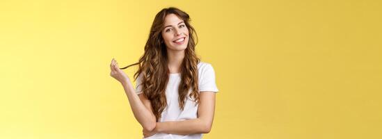 Silly cute feminine sensual curly-haired woman tilt head playing hair strand rolling curl smiling delighted interested listening flirty conversation coquettish gazing camera stand yellow background photo