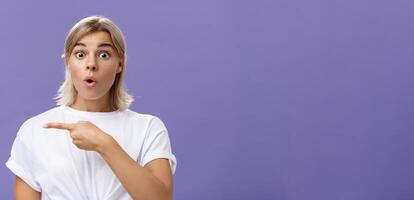 Waist-up shot of overwhelmed excited and surprised attractive urban female in white t-shirt folding lips gasping from amazement being questioned asking question and pointing left over purple wall photo