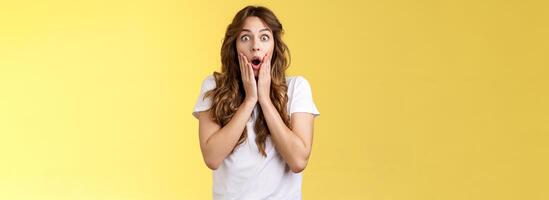 Shocked speechless impressed curly-haired silly girl drop jaw gasping astonished grab cheeks stare camera interested surprised checking out incredible unexpected scene yellow background photo