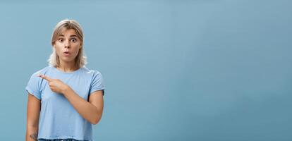 Studio shot of amazed stunned cute blonde witnessing unbelievable event gasping opening mouth staring astonished and pointing left being questioned and shocked over blue background photo