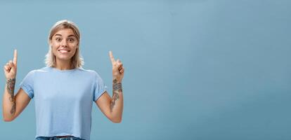 cintura para arriba Disparo de emocionado y emocionado contento maravilloso rubia con corto peinado y tatuajes en brazos sonriente alegremente desde asombro señalando arriba con elevado índice dedos posando terminado azul antecedentes foto