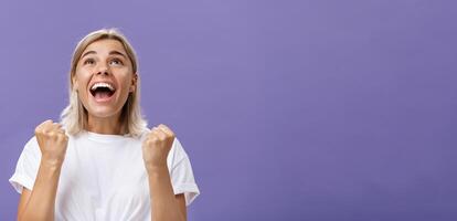 Waist-up shot of grateful delighted lucky girl with attractive tan in white t-shirt clenching fist from positive emotions gazing delighted and happy up thankful giving thanks god for fulfilling dream photo