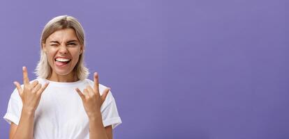 voy a roca. retrato de alegre contento guapo elegante mujer con rubio medio Corte de pelo parpadeo sonriente y pega fuera lengua mientras demostración rock and roll gesto con ambos manos terminado púrpura pared foto