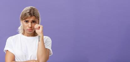 Studio shot of offended sad and timid silly woman with blond hairstyle frowning looking from under forehead holding fist near eye as if whiping teardrop being upset over purple background photo