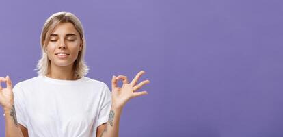 Peaceful calm good-looking female blonde in white t-shirt closing eyes smiling relieved and happy feeling satisfied with life standing in lotus pose with zen gesture over purple background photo