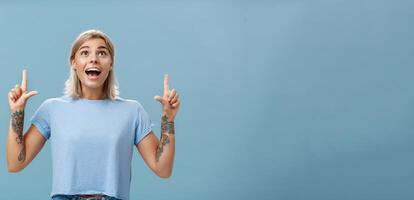 Studio shot of impressed speechless happy good-looking woman with tattoos on arms dropping jaw from amazement and joy gazing fascinated and pointing up standing over blue background photo